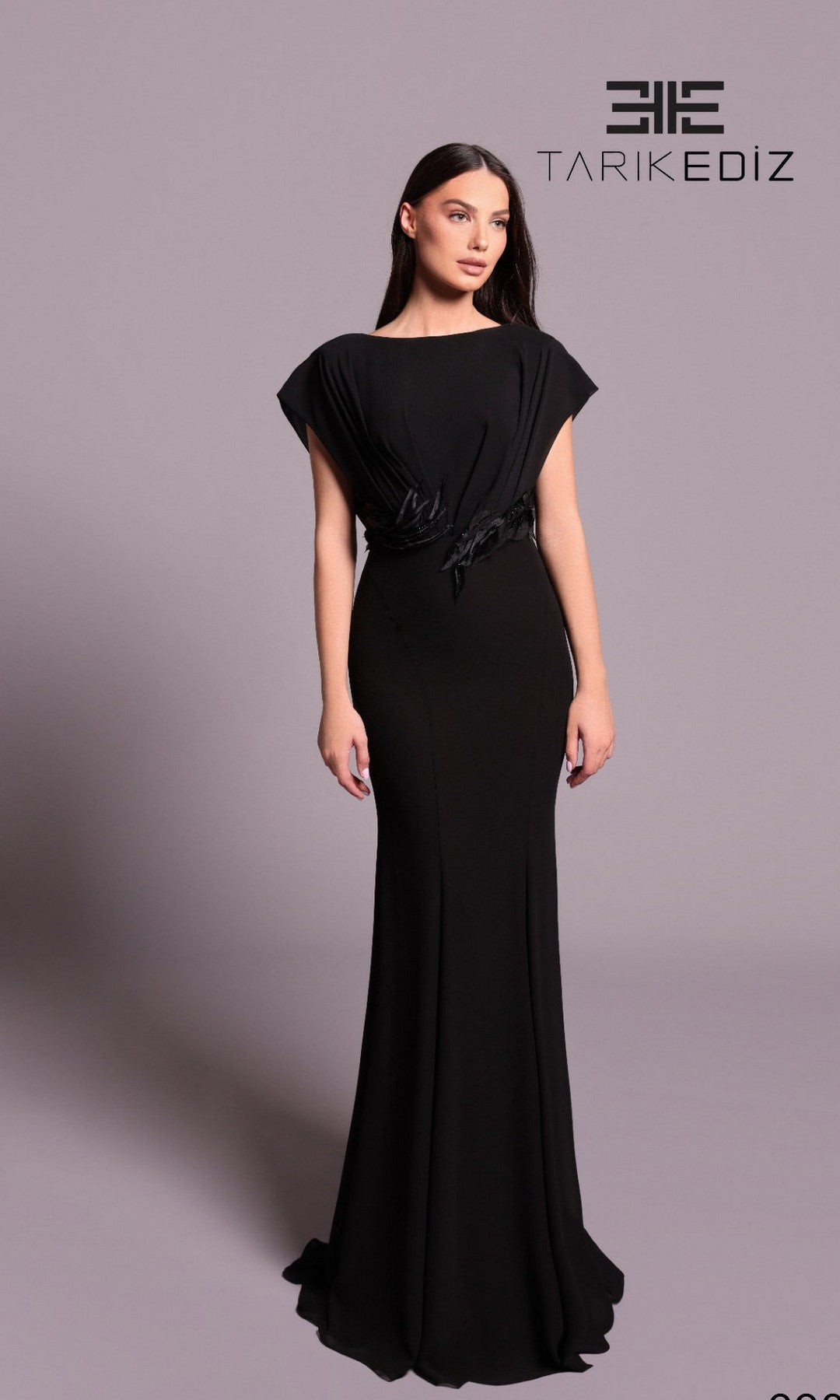 A woman poses against a plain light gray background in the Tarik Ediz 99004—a fitted, floor-length black formal dress with short sleeves and subtle waist draping for a graceful look.